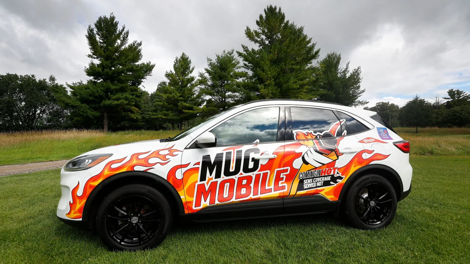 The Mugmobile, a white SUV wrapped in orange flames with Mug Mobile lettering and the Coming In Hot logo, parked side-on on the grass at a golf course.